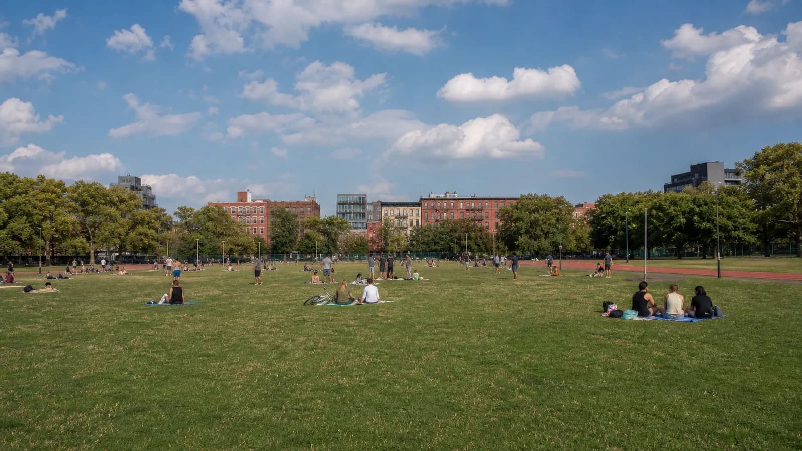 Wide green lawn and running track at McCarren Park with North Brooklyn buildings beyond